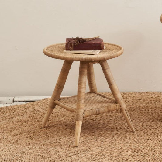 A round woven rattan side table with three twisted legs and a lower support rail, shown on a textured jute rug against a plain beige wall. A small stack of books sits on the tabletop.